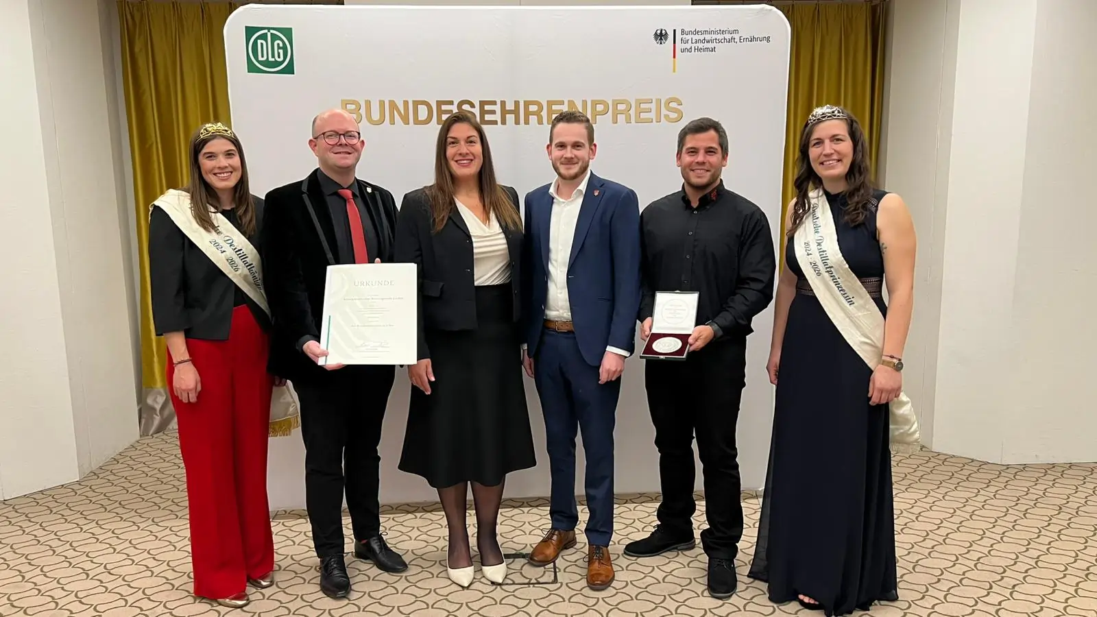 Klosterbrennerei Wöltingerode beim Bundesehrenpreis. 6 Personen vor einer Wand auf der DLG Bundesehrenpreis steht vom Bundesministerim für Landwirtschaft und Ernährung. Von der Klosterbrennerei, Robert Reznizak, Wassiliki Strohmeier, Philipp Becker, Dany Mailah.